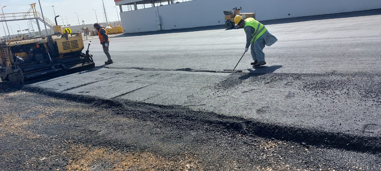 Paver and crew laying asphalt while a worker rakes the edge of the mat